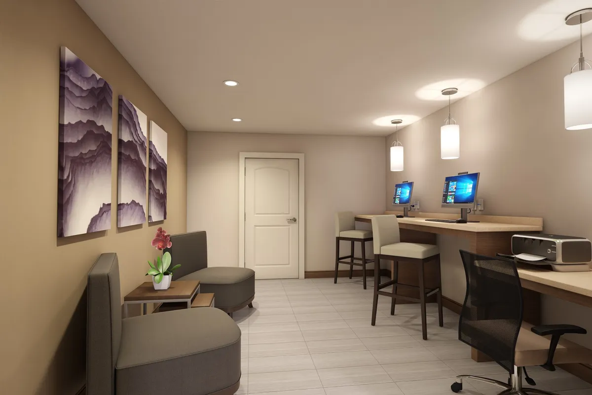 This image depicts a contemporary business center featuring computer stations, a seating area adorned with plants, and abstract mountain-themed artwork. The environment is bright and modern, inviting both productivity and relaxation, with several workstations available and a communal seating area nearby.