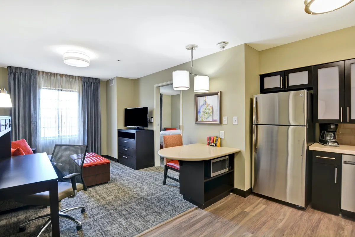 The image highlights a compact but fully functional living area with a sofa, TV, work desk, and a kitchenette in the background. The combination of workspace and kitchen area, along with relaxing seating, demonstrates the suite's versatility for both business and leisure stays.