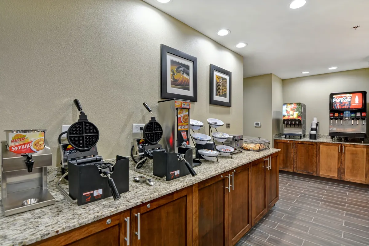 A welcoming breakfast area with granite countertops, self-serve waffle makers, syrup and drink dispensers, and framed artwork on the wall. Guests are seen making waffles and enjoying breakfast together, creating a sense of convenience and sociability.