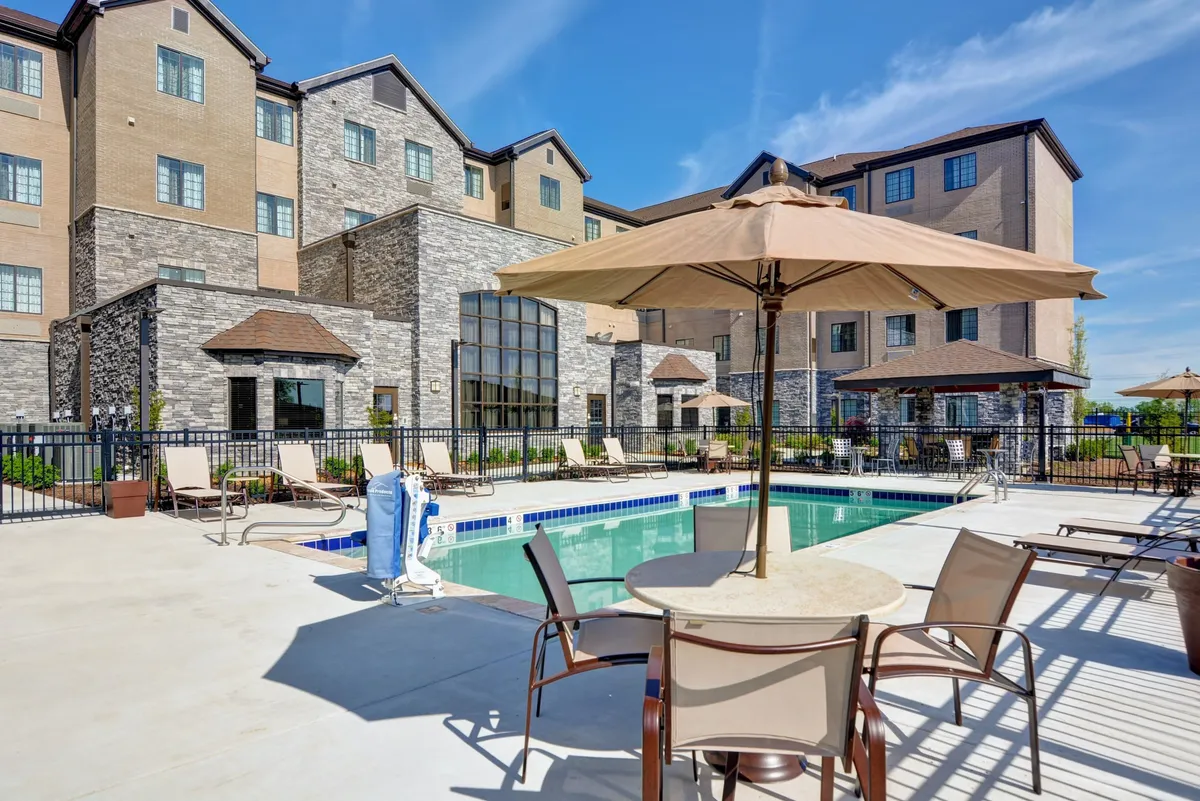 This image captures the outdoor pool area on a sunny day, featuring inviting lounge chairs and umbrellas arranged around the sparkling pool. The building's facade frames the scene, while clusters of guests relax, sunbathe, and socialize, evoking the leisurely atmosphere of pool season.