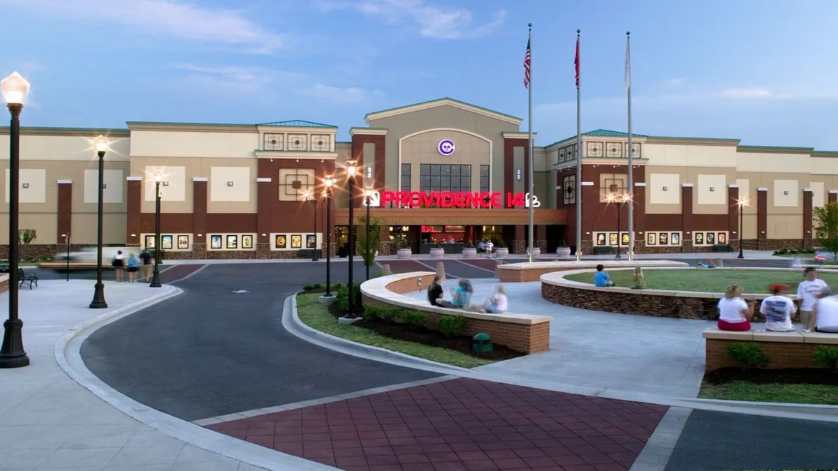 The exterior of a bustling local theater, with bright signage and an animated plaza filled with seating and decorative lampposts. The scene hints at community life and the proximity to entertainment and cultural events.