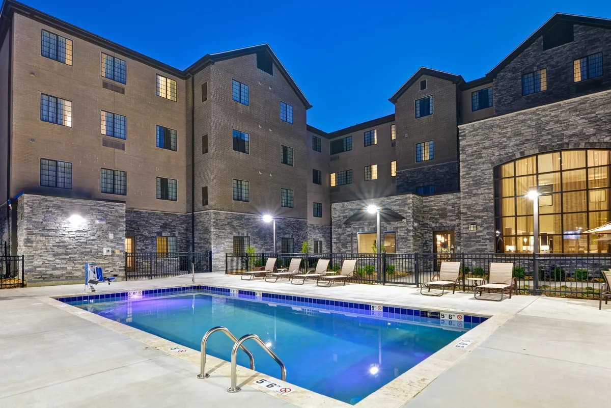A vibrant exterior view of the property featuring the swimming pool, lounge chairs arranged for relaxation, and the building's facade. The scene is bathed in natural light, creating a welcoming atmosphere that suggests leisure as well as a break between business activities.