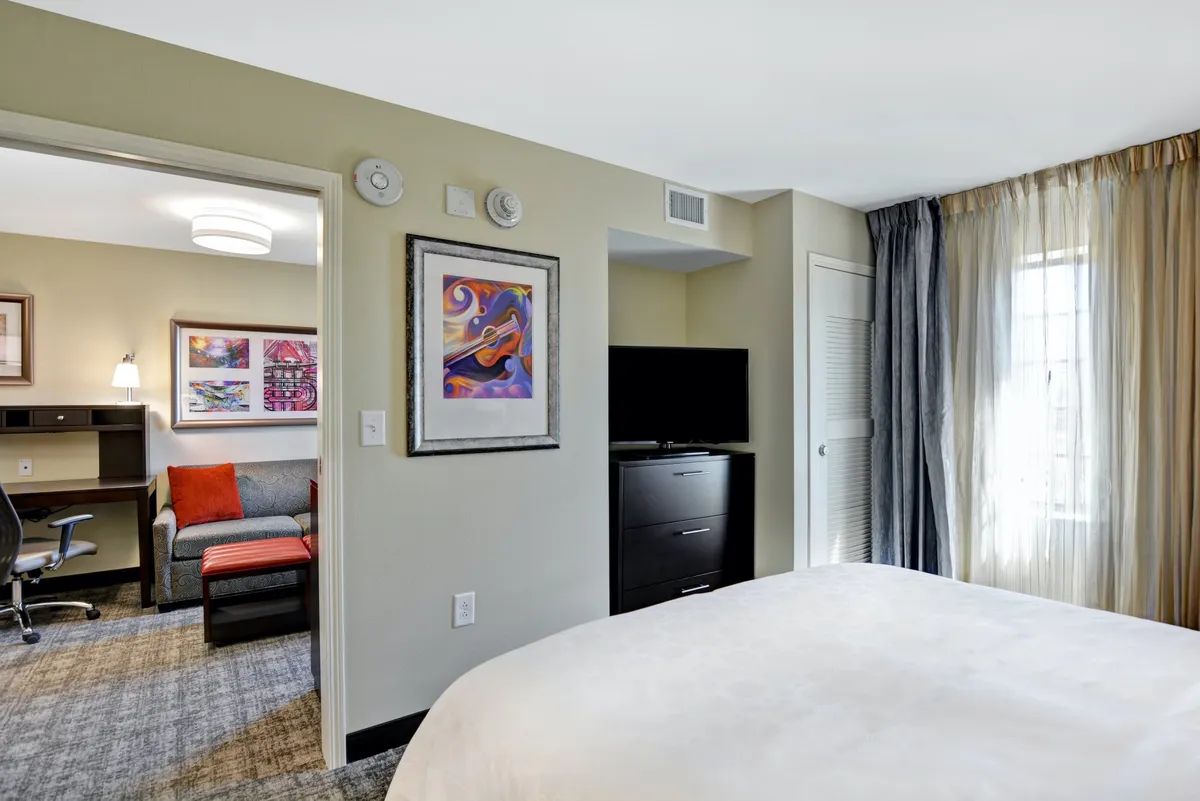 This interior image shows a cozy suite featuring a plush king bed, a stylish sofa, a dedicated desk area, and a mounted television. The arrangement highlights separation between the bedroom and living space, with tasteful wall art and a calm, restful ambiance. The desk area suggests suitability for business travelers.