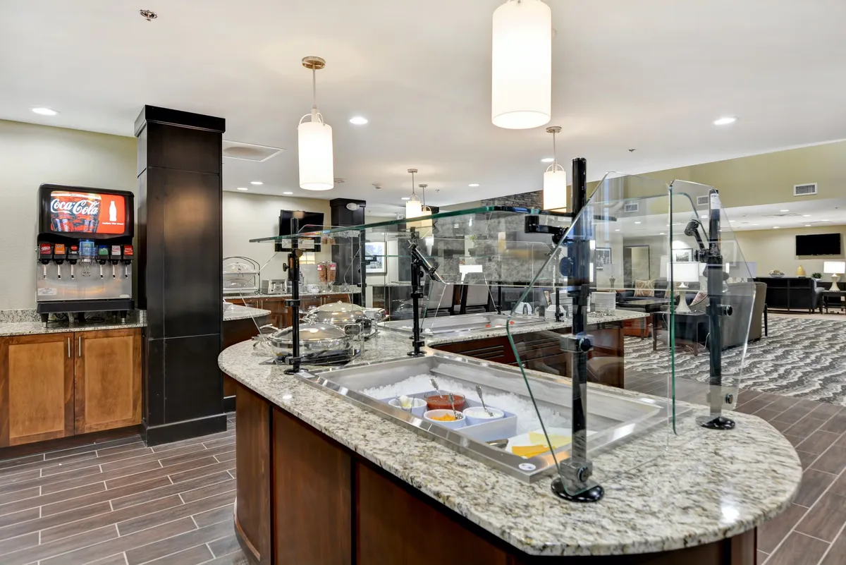 A bright and casual food display area with a granite countertop, beverage dispenser, and hanging light fixtures, capturing the lively atmosphere of guests enjoying breakfast or a quick meal together.