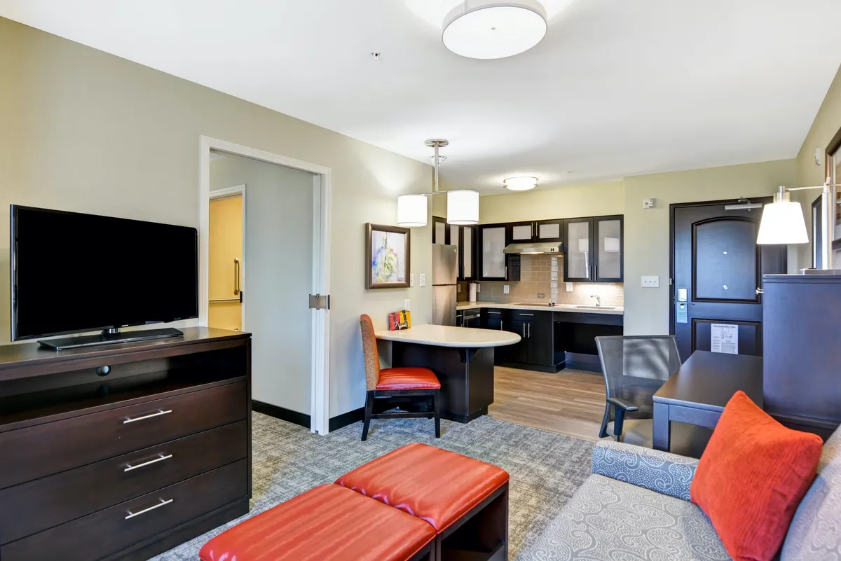 The image showcases a bright, welcoming condo interior featuring a comfortable sofa and armchair arranged around a large TV, a small dining table perfect for two, and a fully equipped kitchen area. The space is styled for relaxation and convenience, suggesting easy nights in after exploring nearby attractions.