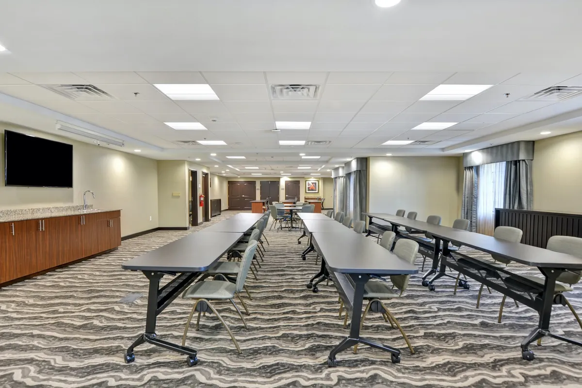 This image reveals a meeting room with long tables organized in a U-shape, a television screen mounted on the wall, large windows letting in natural light, and sleek wooden cabinetry. The setup is conducive for team meetings, workshops, and professional presentations, evoking a collaborative atmosphere.